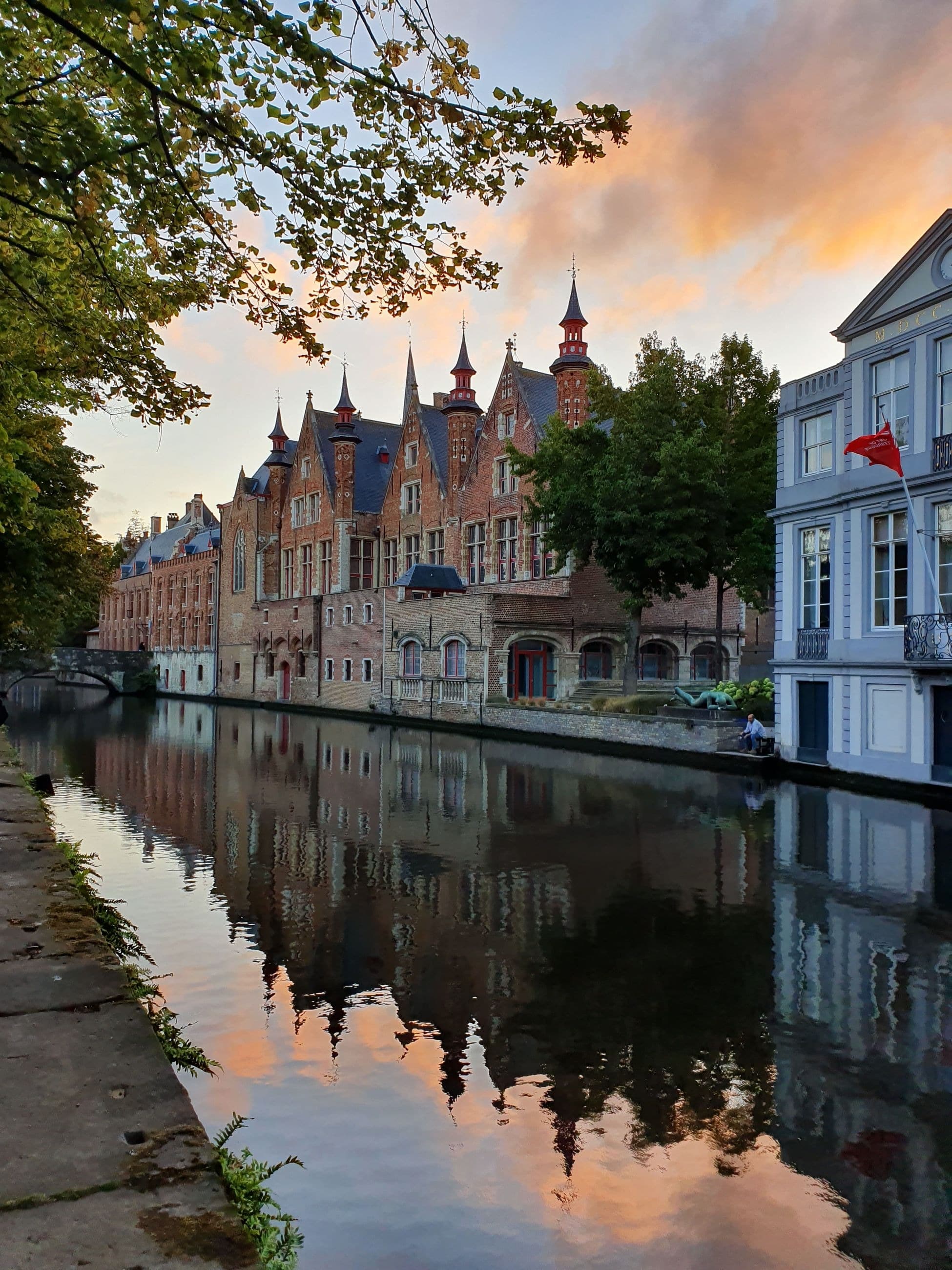 A row of Gothic stepped-gable buildings reflected in a still Bruges canal at dusk, under warm orange clouds