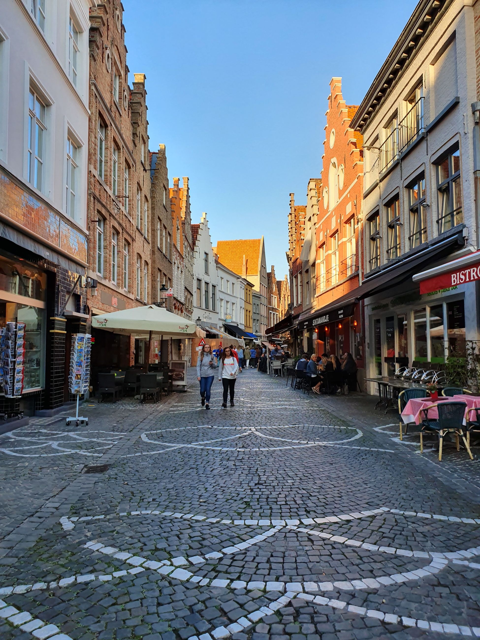 A quiet cobblestone street in golden evening light, with cafe terraces and old gabled houses on either side
