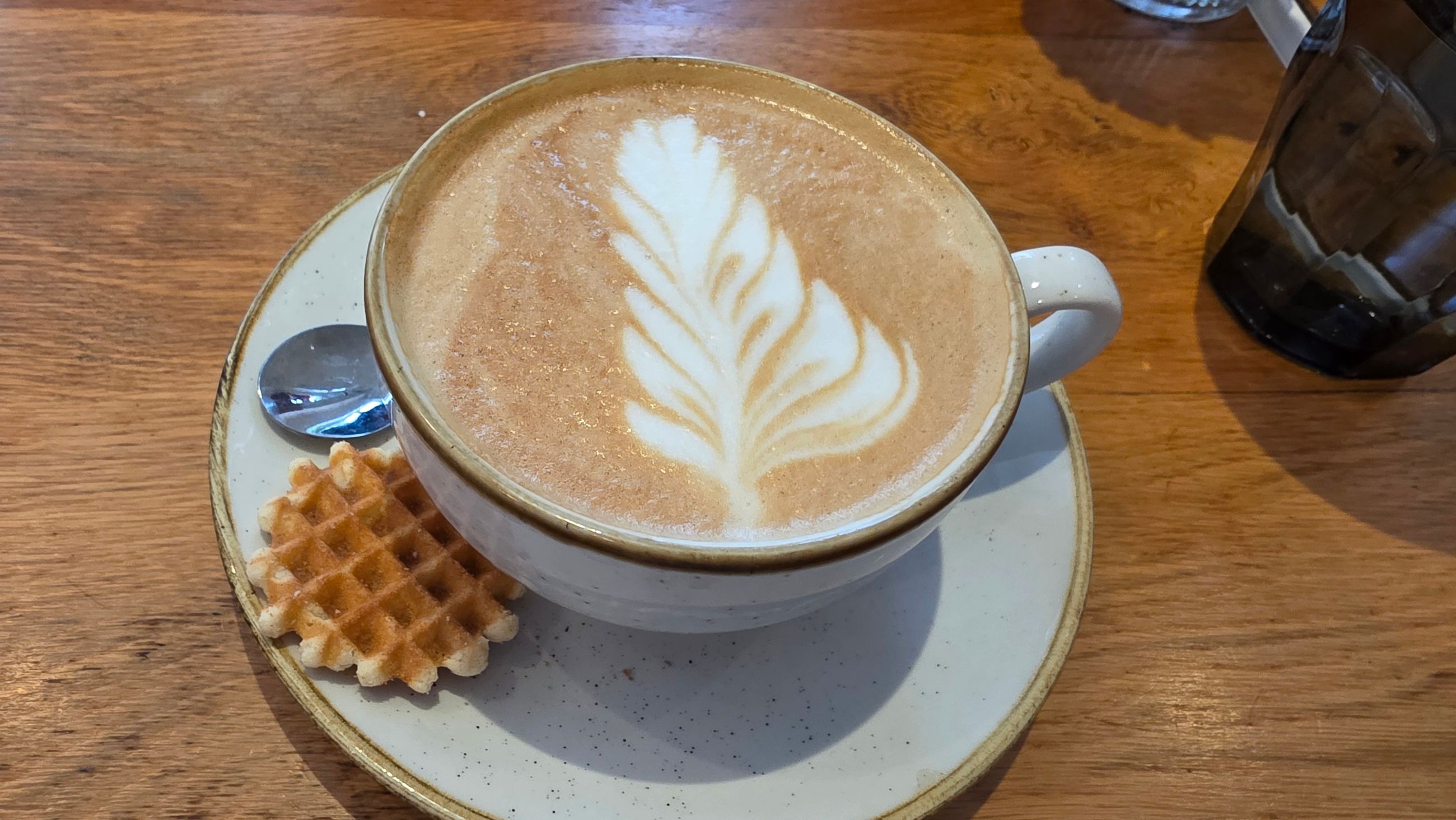A cappuccino with leaf-pattern latte art and a mini waffle on the saucer, at That's Toast